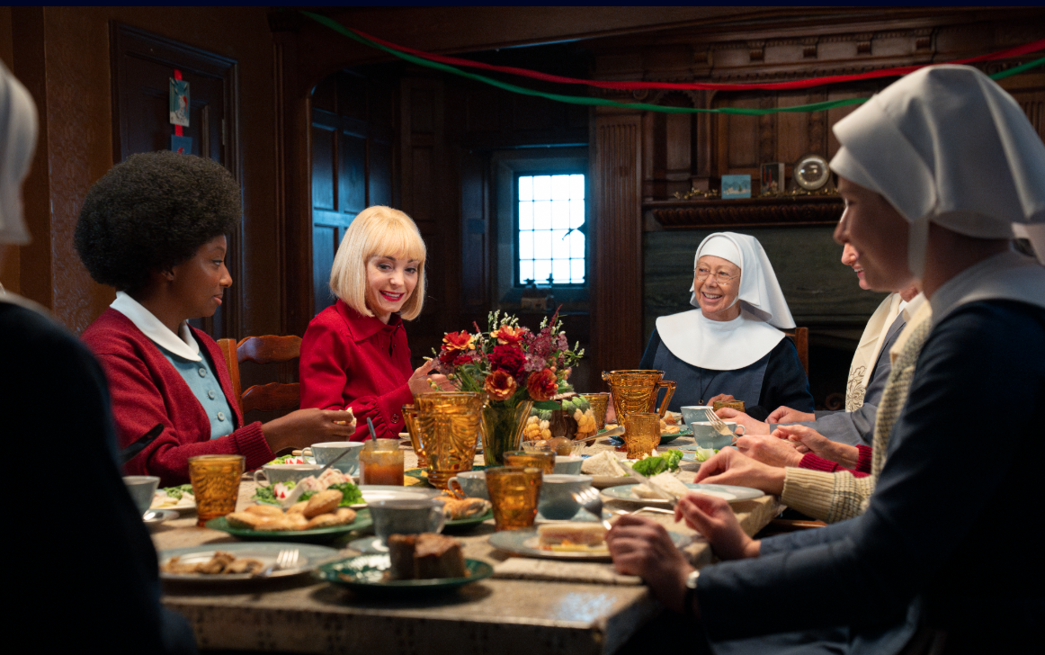 Four nuns and two nurses sit around a table eating a meal together.